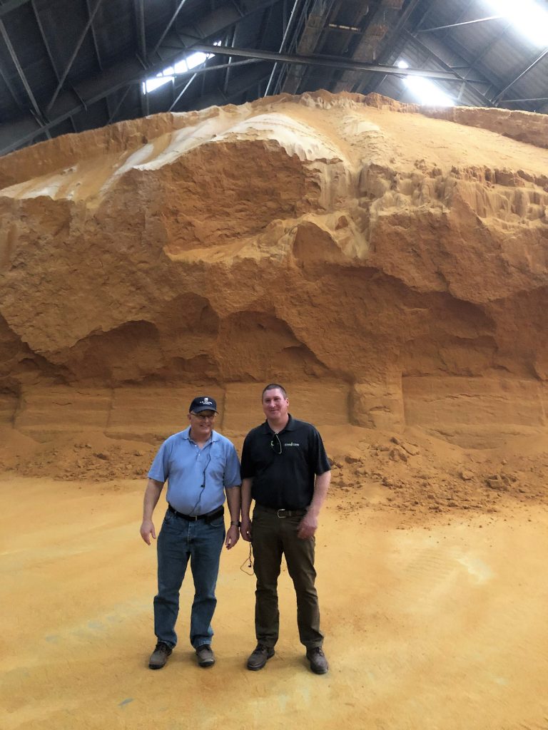 Two Men Standing In front of a large pile of DDGS