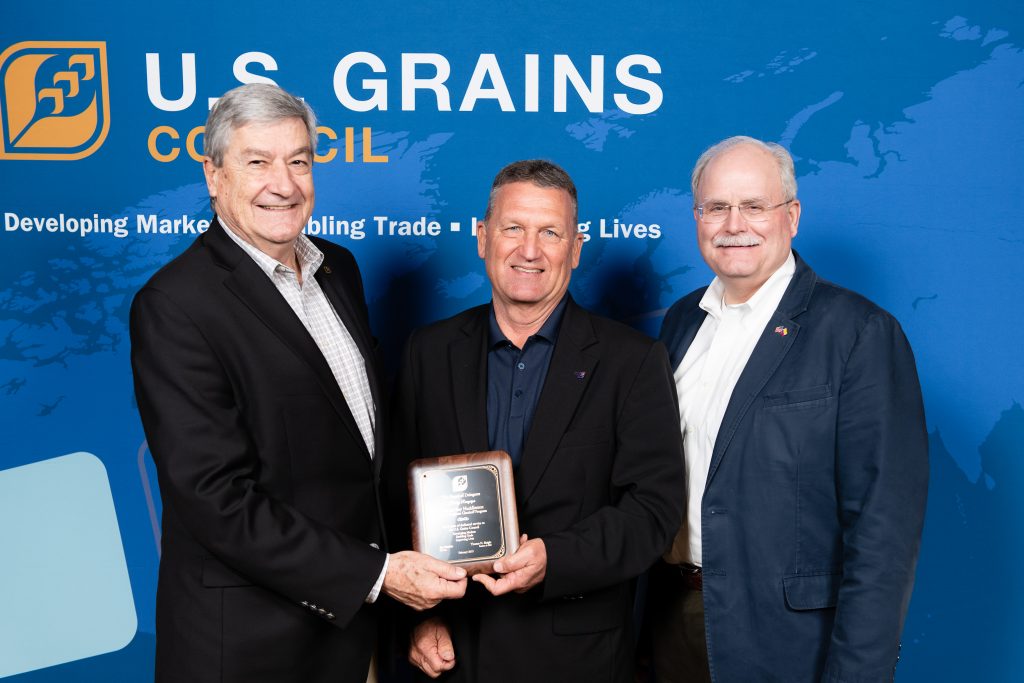 Charles Ray Huddleston Recognition, 3 men on stage, man in the middle holding a recognition plaque