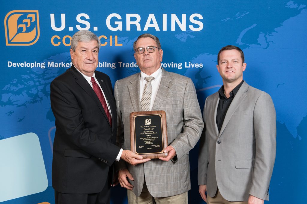 David Gibson Recognition- 3 men standing in front of the USGC banner, man in the middle holding recognition plaque