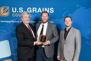 3 men standing in front of the USGC banner, man in the center holding a recognition plaque