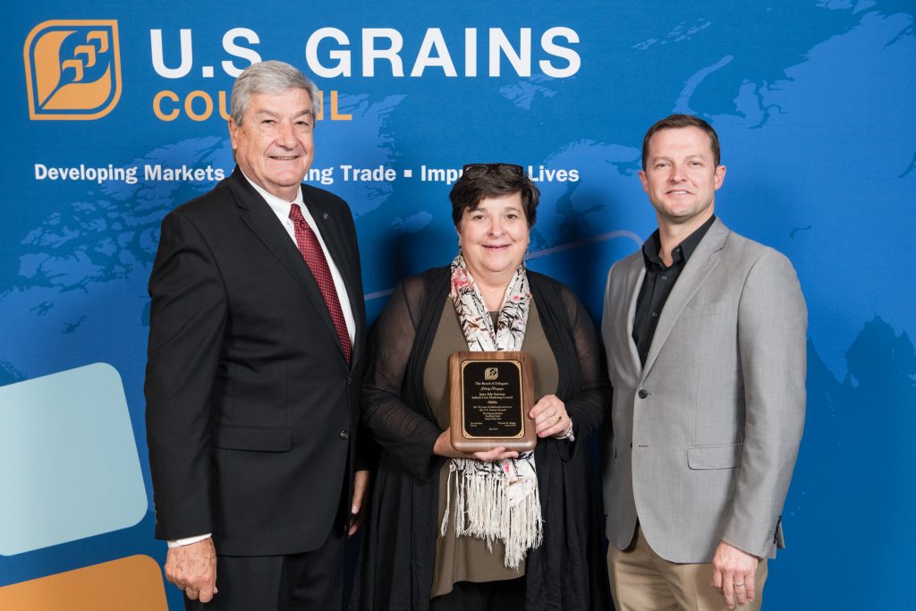 Jane Ade Stevens Recognition- 2 men & 1 woman standing in front of the USGC banner, woman in the middle holding recognition plaque