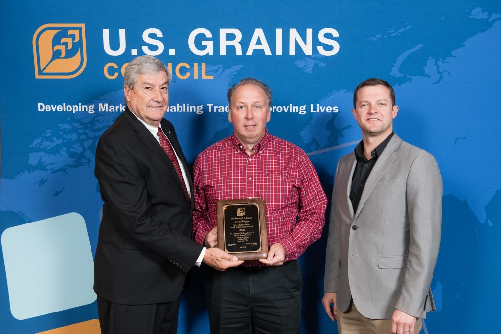 Steve Edwardson Recognition- 3 men standing in front of the Grains Council backdrop, man in the middle holding a recognition plaque