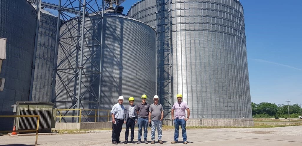 Five Guys in Hard Hats Standing In Front Of Large Metal Silos