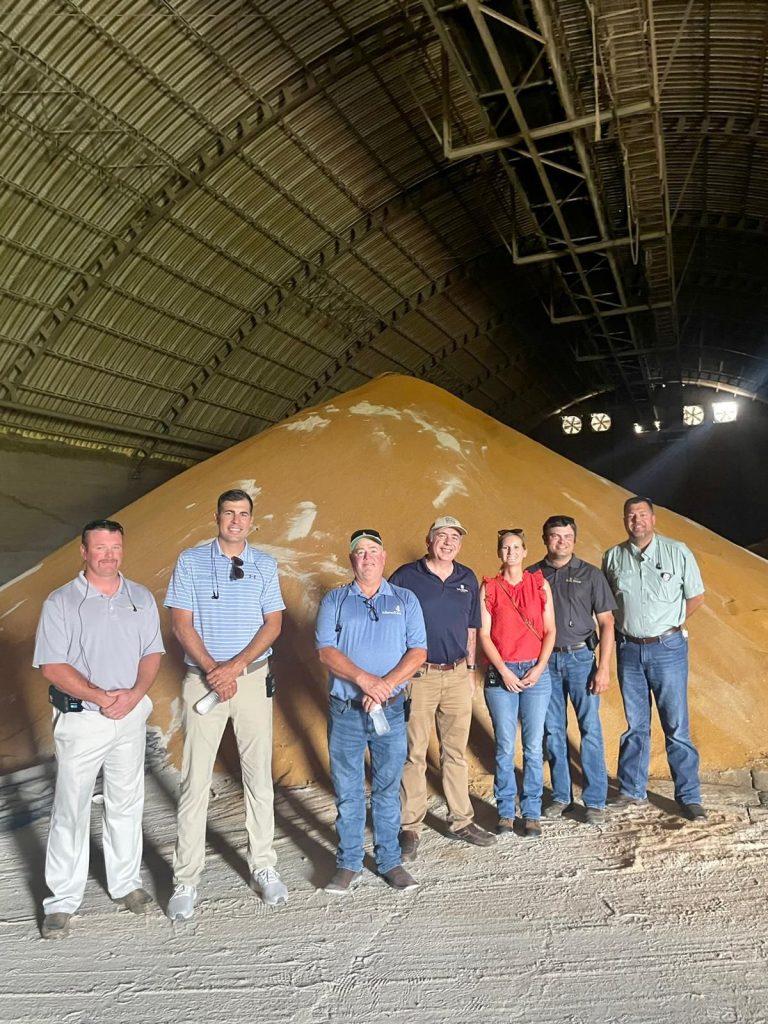 group of people smiling for a photo in front of a pile of DDGS