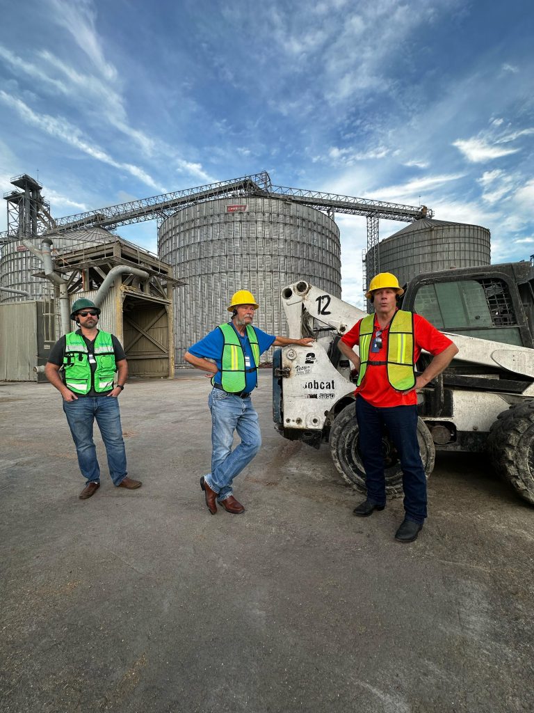 Three men in safety gear stand in front of a grain storage facility