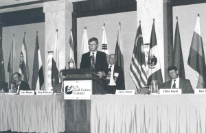 A black and white photo of a man at a podium in front of various national flags