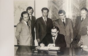 A man signing a document with four men observing